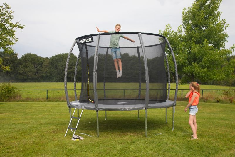 Zwei Kinder nutzen das KETTLER Trampolin JUMP 305 im Garten. Ein Kind springt hoch, während das andere zuschaut.