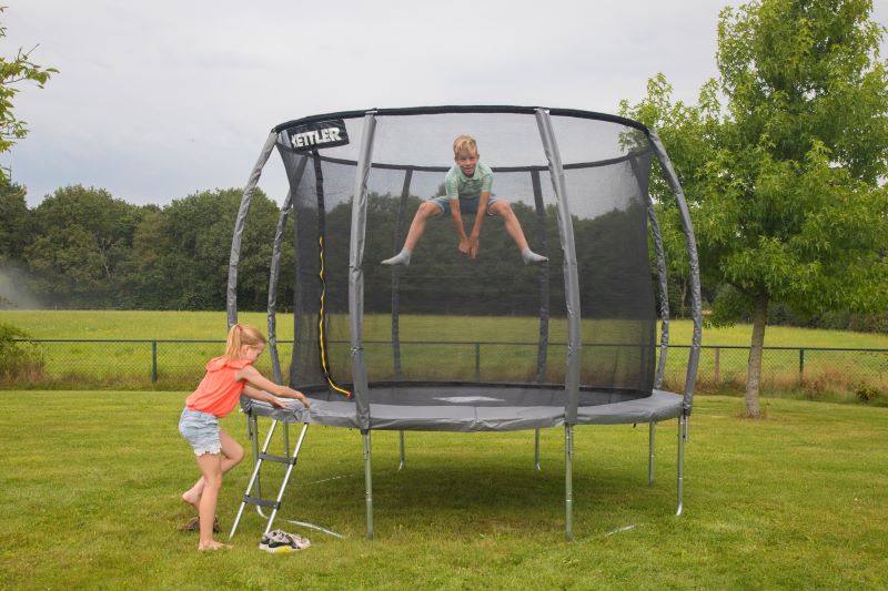 Ein Kind springt auf dem KETTLER Trampolin JUMP 305 im Garten, während ein weiteres Kind die Leiter hochsteigt.